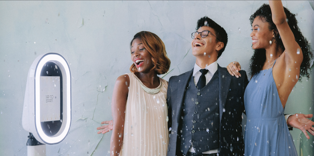 Three wedding guests laugh under falling confetti while posing beside a modern ring-light photo booth.