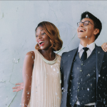Three wedding guests laugh under falling confetti while posing beside a modern ring-light photo booth.