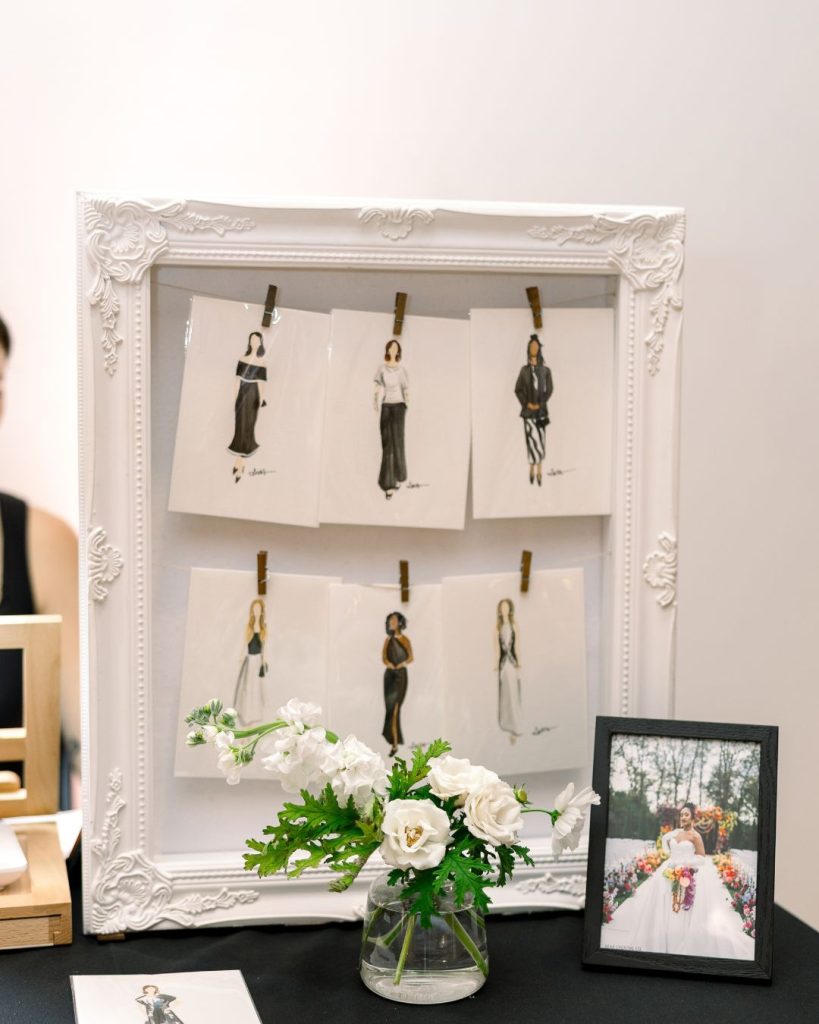 Illustrated guest portraits clipped inside an ornate white frame beside a vase of white flowers.