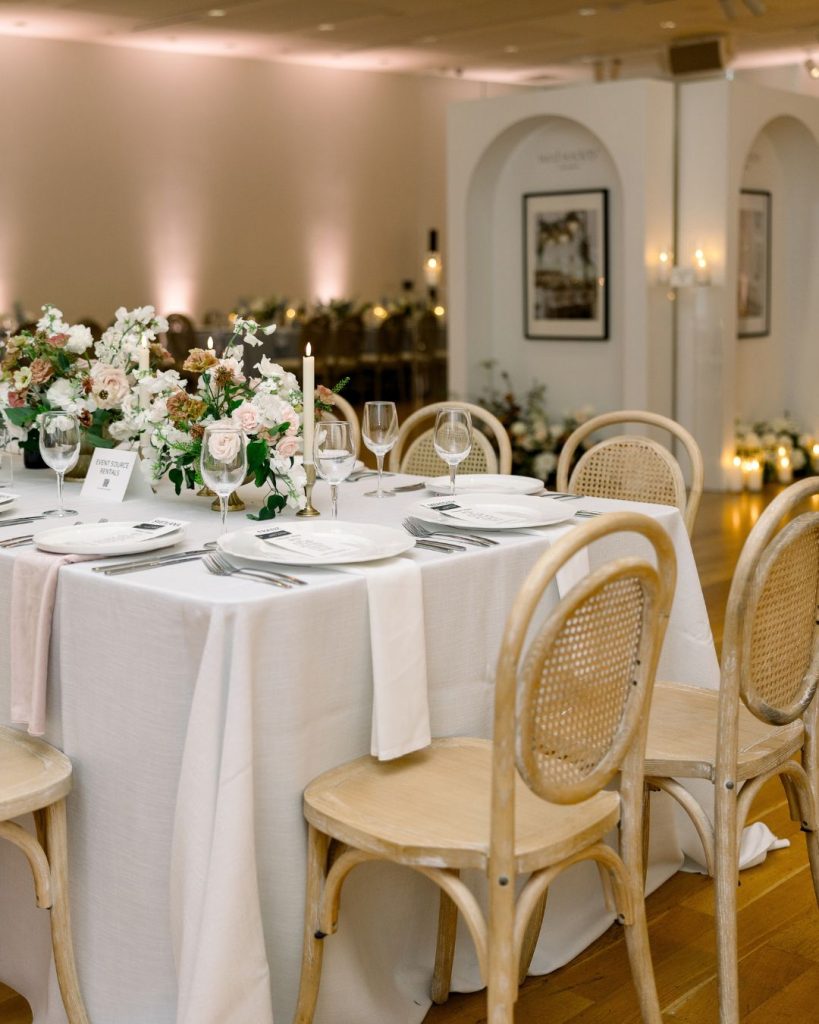 Long reception table with blush linens, pastel floral centerpieces, white plates, and cane back chairs.