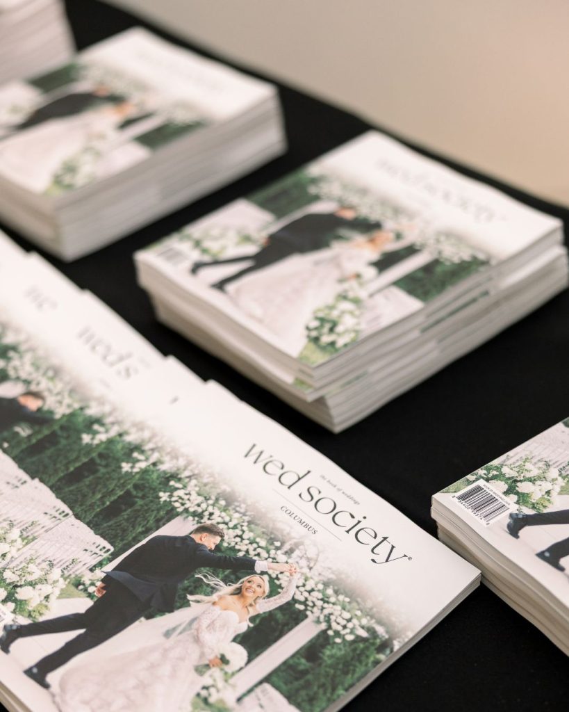 Stacks of the inaugural 2026 Wed Society Columbus Book of Weddings on a black display table.