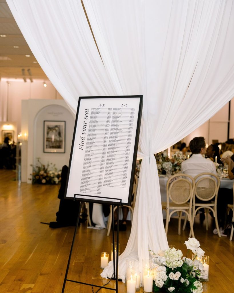 Framed “Find your seat” seating chart beside flowing white draping, floral arrangements, and pillar candles.