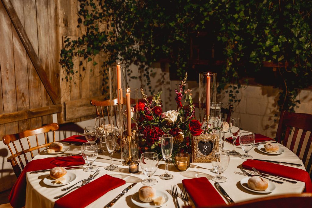 Round table set with red napkins, tall pink candles in glass cylinders, and a lush red-and-white floral centerpiece with table number 8.