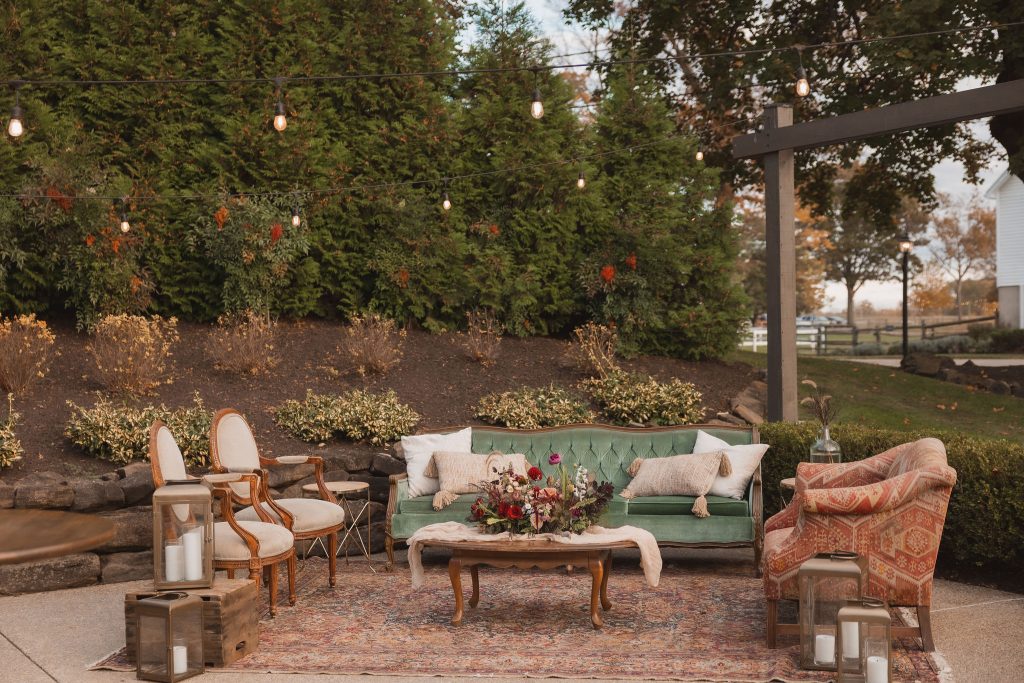 Outdoor lounge area with a green velvet sofa, patterned armchair, wooden chairs, and floral arrangement under string lights.