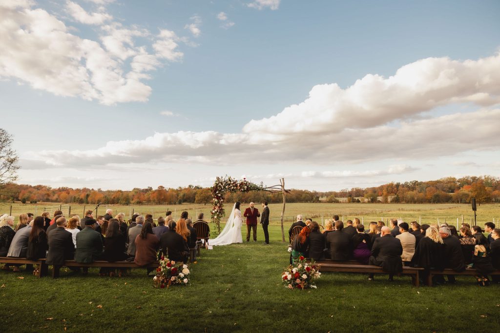 Bride and groom stand under the floral arch with the officiant during their outdoor ceremony as guests watch from wooden seats.