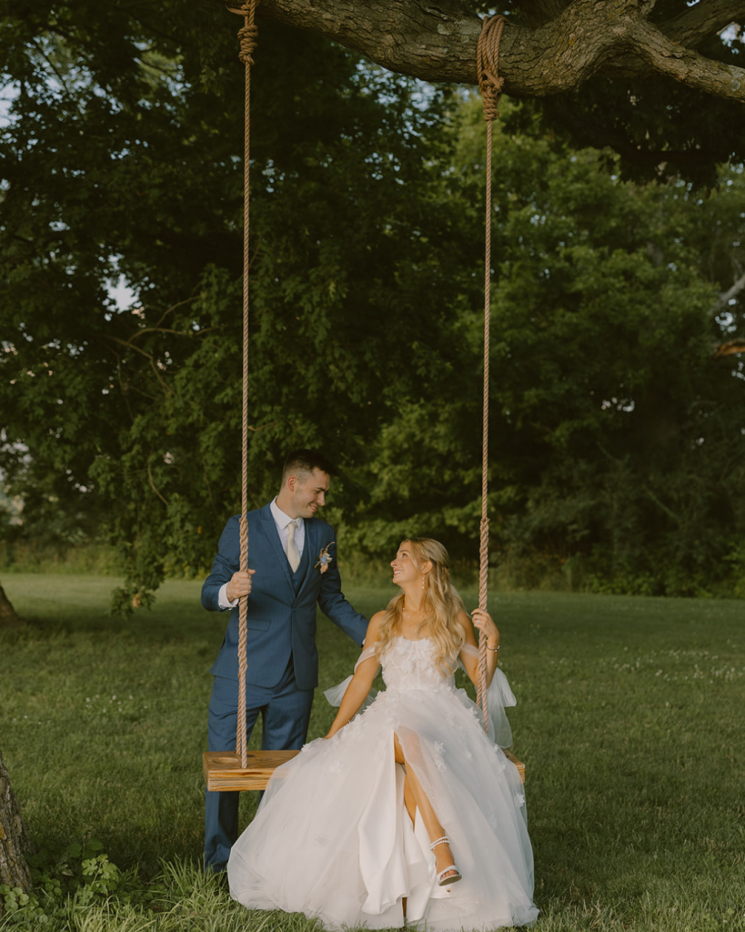 Groom and bride sit together on a wooden swing outdoors during their summer wedding.