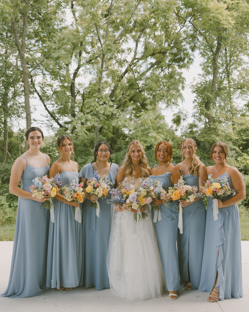 Bridesmaids in blue dresses pose outdoors with colorful floral bouquets.