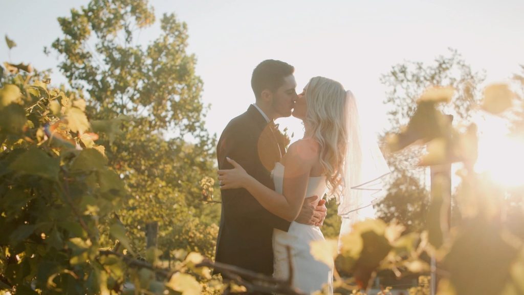 Bride and groom kiss among grapevines at sunset while their Columbus wedding videographer captures the golden-hour moment.