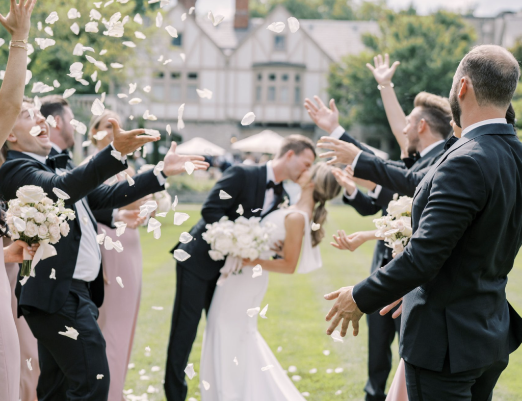Columbus Ohio wedding photographer captures a bride and groom kissing as their wedding party tosses white flower petals during an outdoor celebration, with bridesmaids in blush pink dresses and groomsmen in black tuxedos.