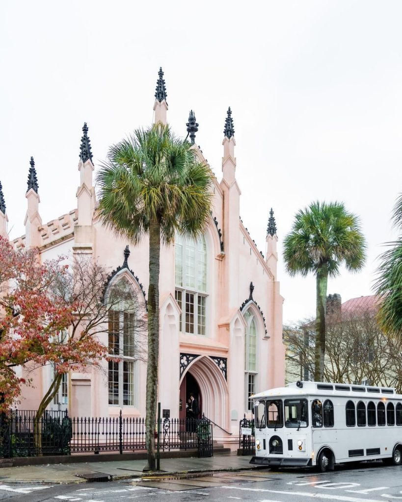 Exterior view of a historic light-pink Gothic Revival church with palm trees in front and a white wedding trolley parked along the street.