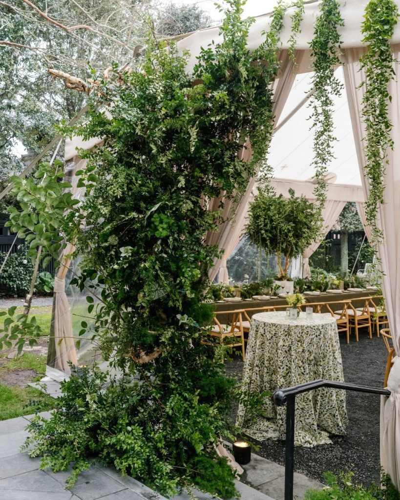 Tented reception entrance framed by hanging greenery and climbing vines, with a sequined tablecloth and long feasting table visible inside.