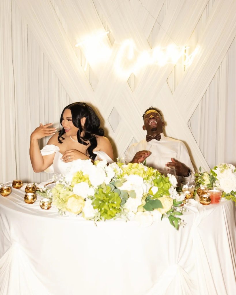 bride and groom laughing together at wedding reception under LED sign and gold drapery sweetheart table