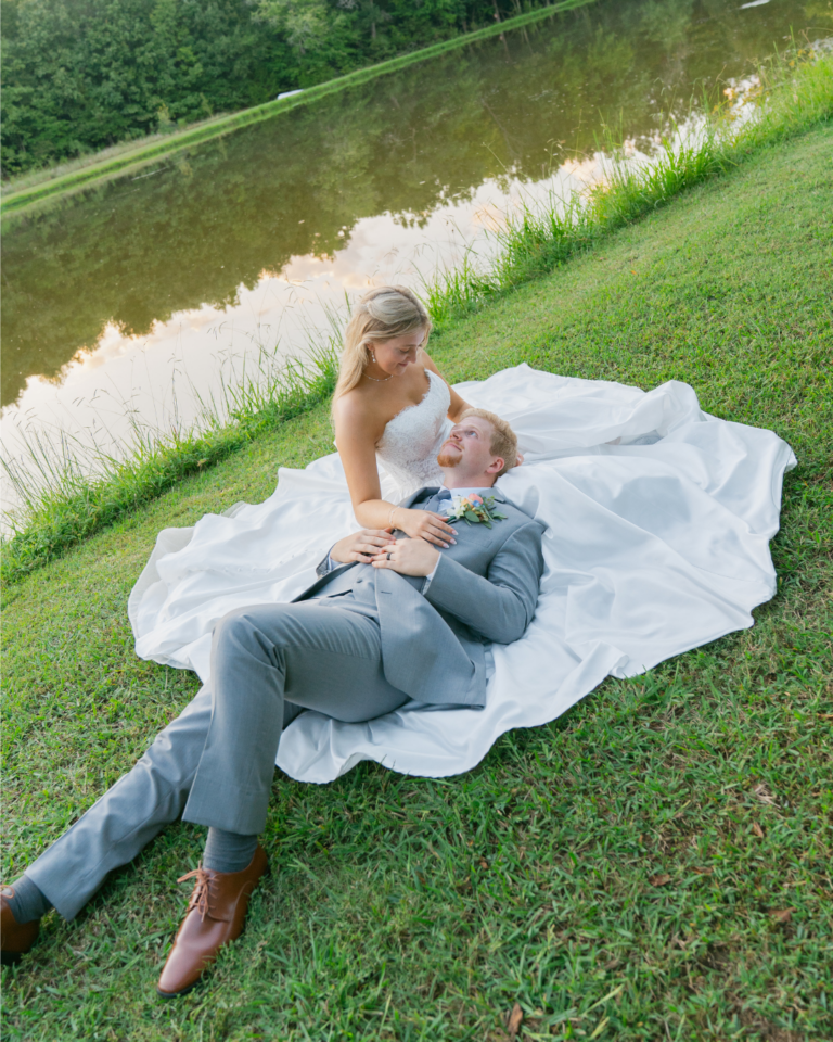 Abby sits on the grass beside a pond at Cedar Hills in Salisbury, NC, with Connor resting in her lap and her white wedding gown spread around them.