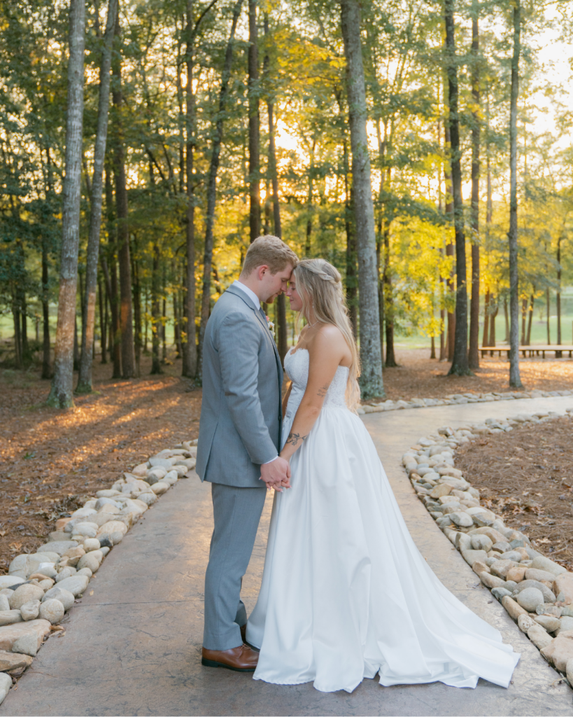 Abby and Connor stand forehead to forehead on a wooded pathway at Cedar Hills, surrounded by trees and warm golden evening light.