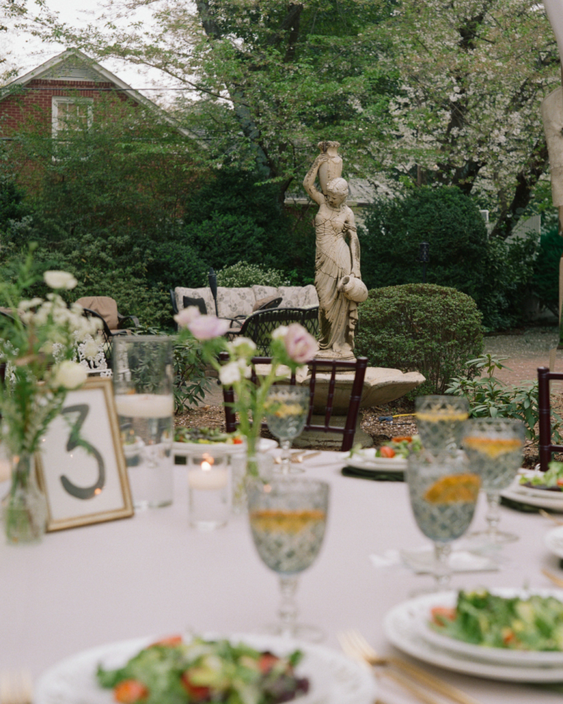 Elegant outdoor wedding reception table at The Morehead Inn with patterned glassware, white linens, candles, and garden surroundings.