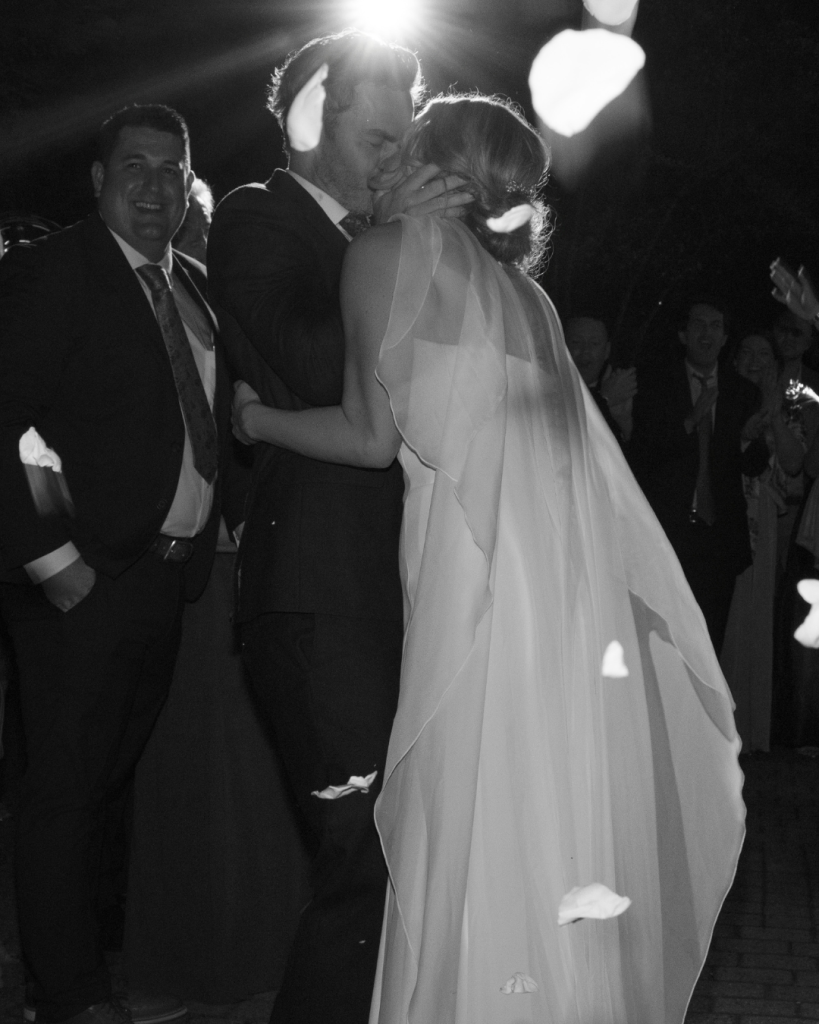 Black and white photograph of Elizabeth and Tom kissing during their wedding celebration as guests look on.