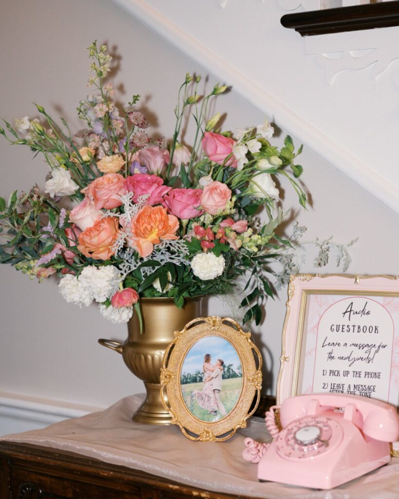A floral centerpiece with pink, peachy, and white roses sitting on a table next to a vintage pink telephone and framed photos.