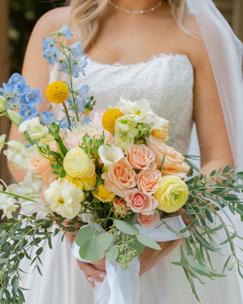 Closeup of Abby's garden inspired bridal bouquet featuring peach roses, yellow ranunculus, blue delphinium, white flowers, and greenery.