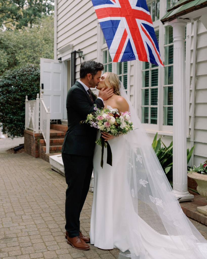 Charlotte wedding couple Elizabeth and Tom kiss beneath a Union Jack flag outside The Morehead Inn, honoring the groom's English heritage.