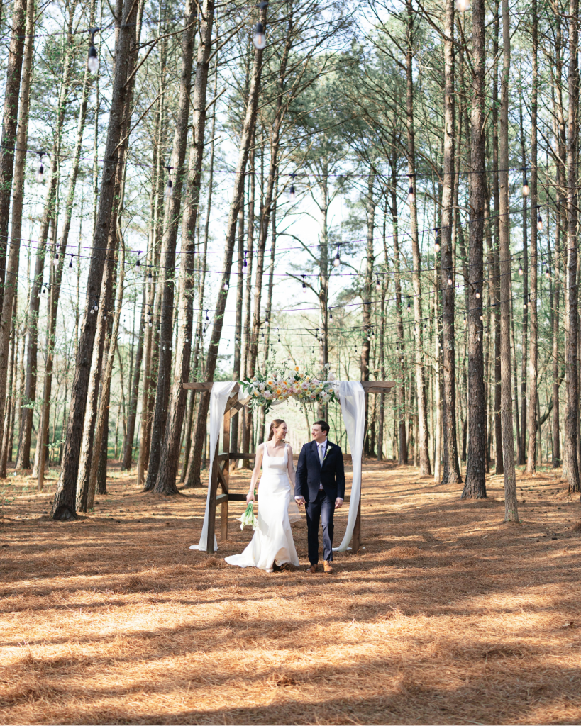 Tate and Alex walk beneath a floral accented wooden ceremony arbor surrounded by tall pine trees at The Union Exchange.