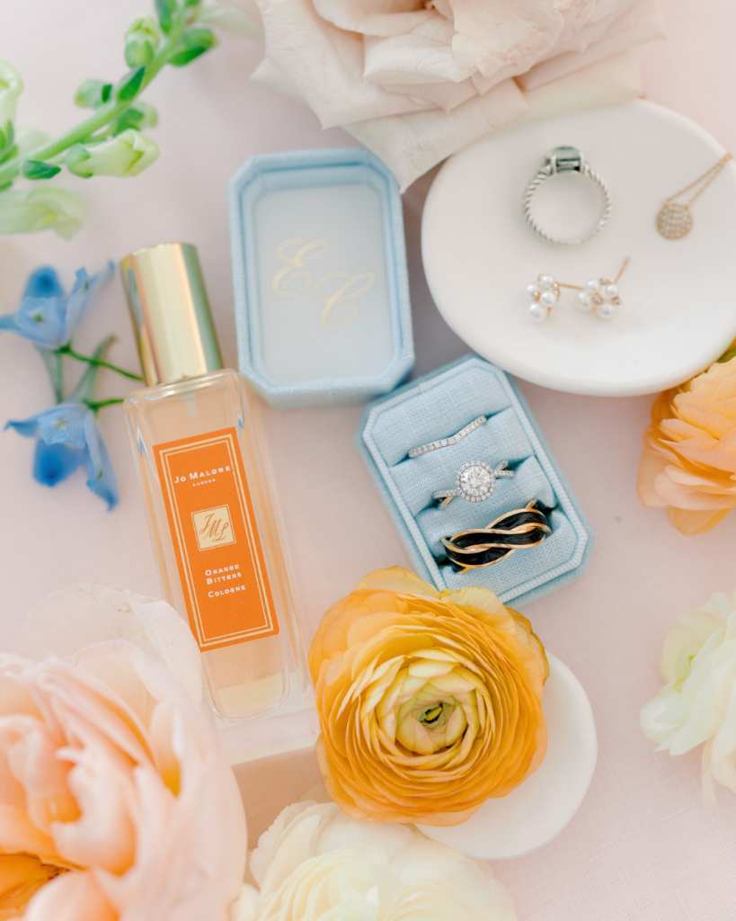 Bridal detail flat lay featuring wedding rings, pearl earrings, Jo Malone perfume and peach, white and blue spring flowers.