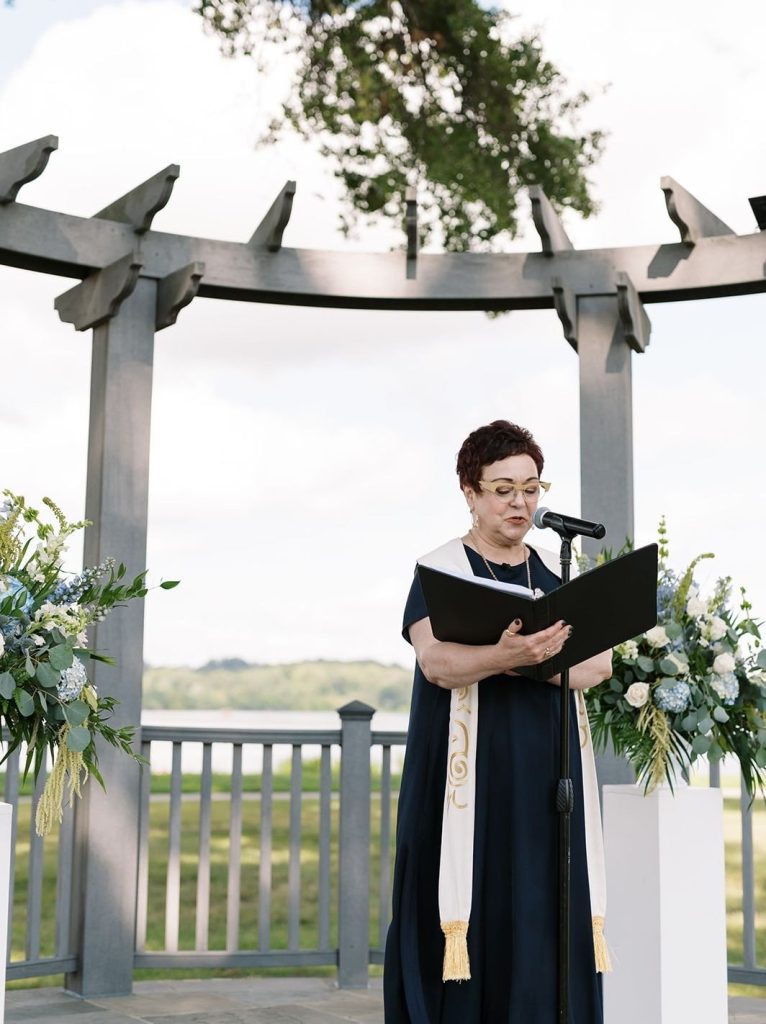 Female wedding officiant standing at a microphone reading from a black binder under a wooden gazebo with a waterfront view.