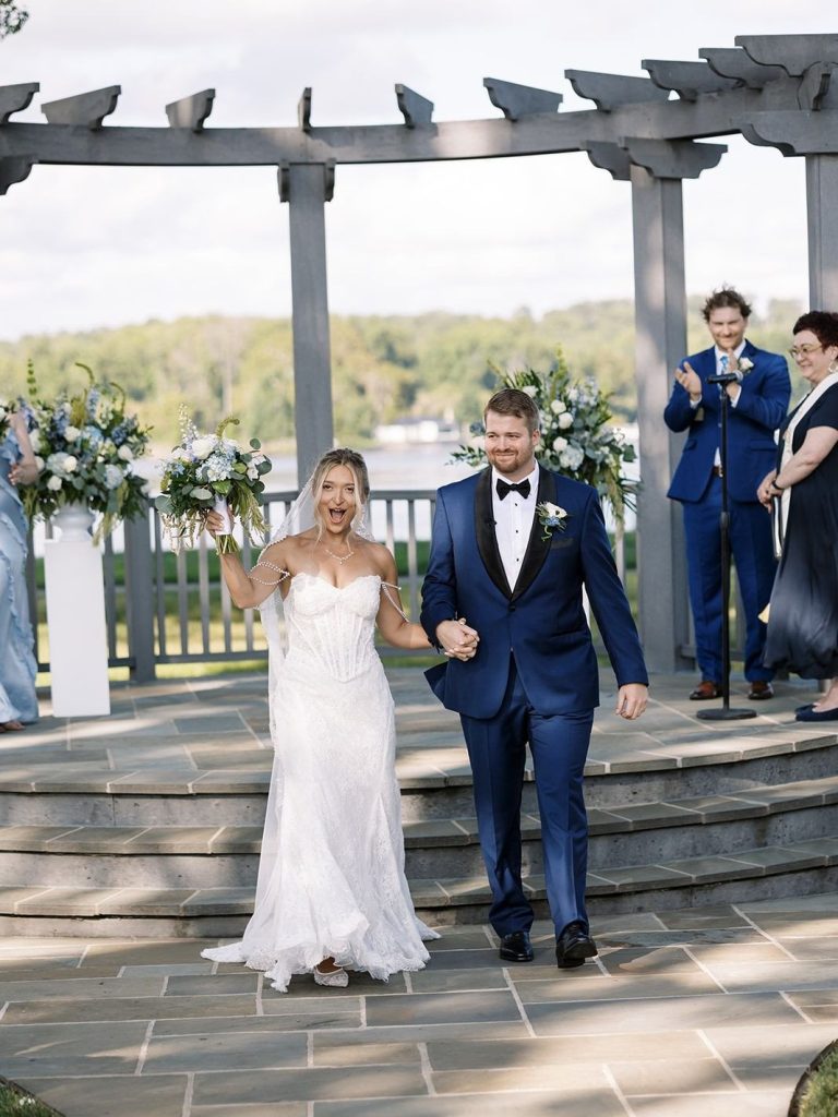 Joyful bride and groom holding hands and cheering as they walk down the steps of a waterfront gazebo following their wedding ceremony, while guests applaud in the background.