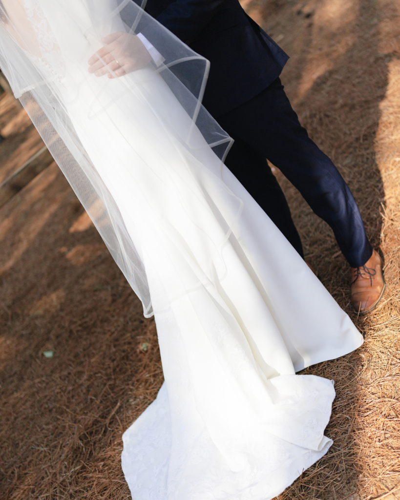 Close detail of Tate’s white wedding gown and veil beside Alex’s navy suit during portraits in the wooded venue setting.