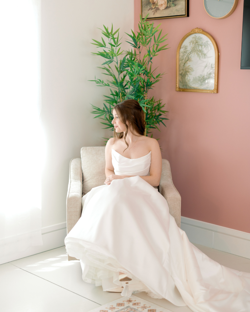 Bride Emma sits beside a window in her strapless white wedding gown before her Splendor Pond wedding ceremony.