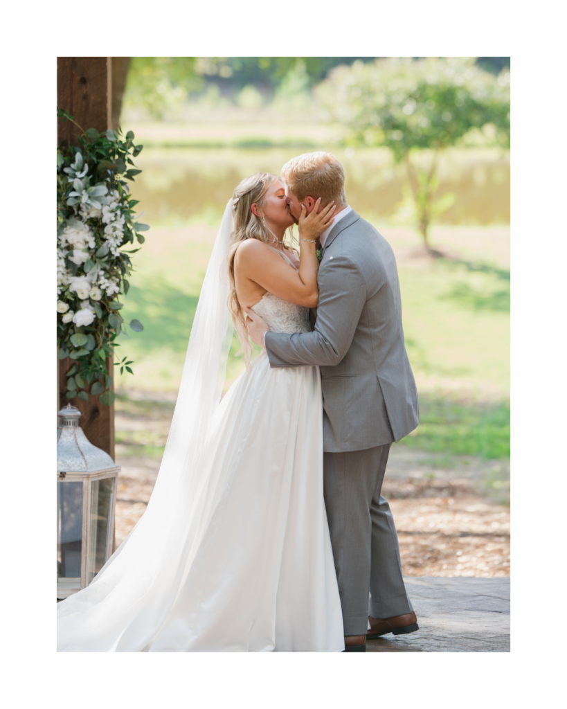 Abby and Connor share a kiss beneath a rustic wooden structure decorated with white flowers and greenery at Cedar Hills.