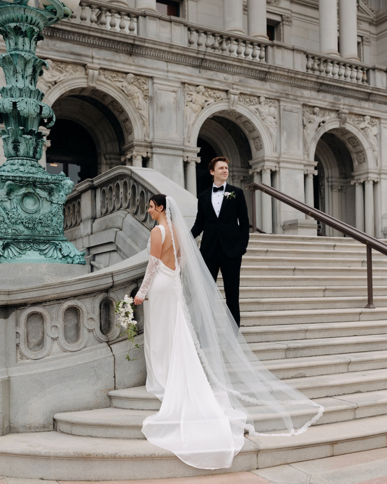 Bride in a flowing white wedding gown and long veil walks down the grand stone steps of a historic building, while the groom in a black tuxedo stands behind her beneath ornate archways.