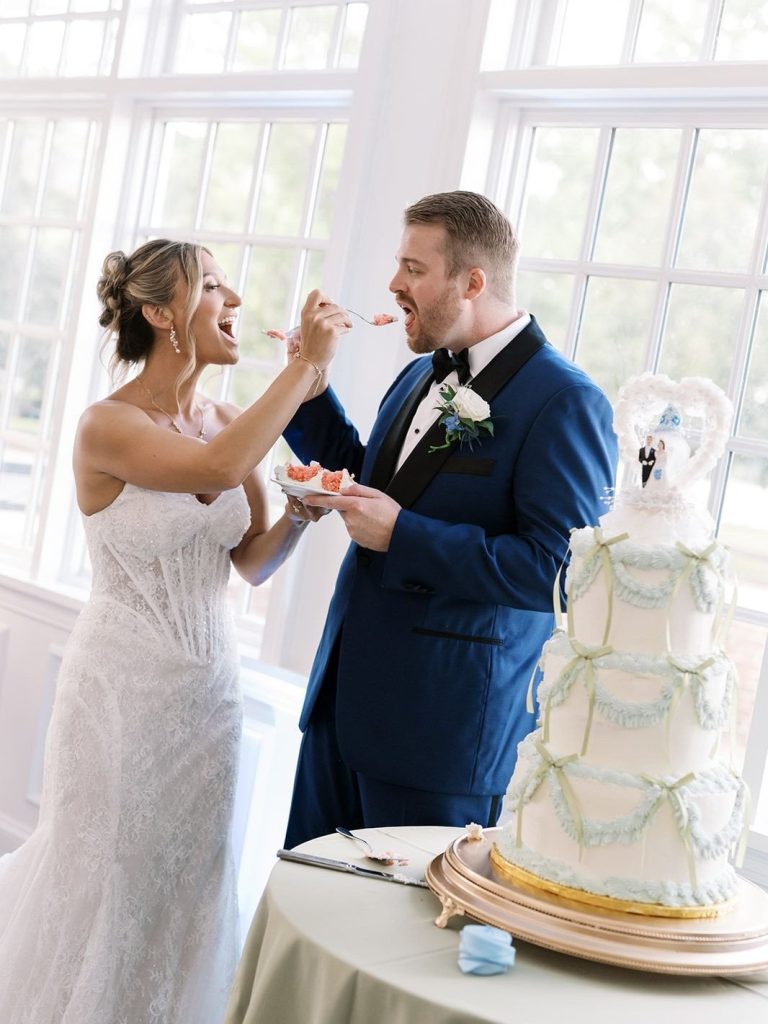 Bride playfully feeding a bite of wedding cake to the groom while standing indoors next to their three-tiered cake and large windows.