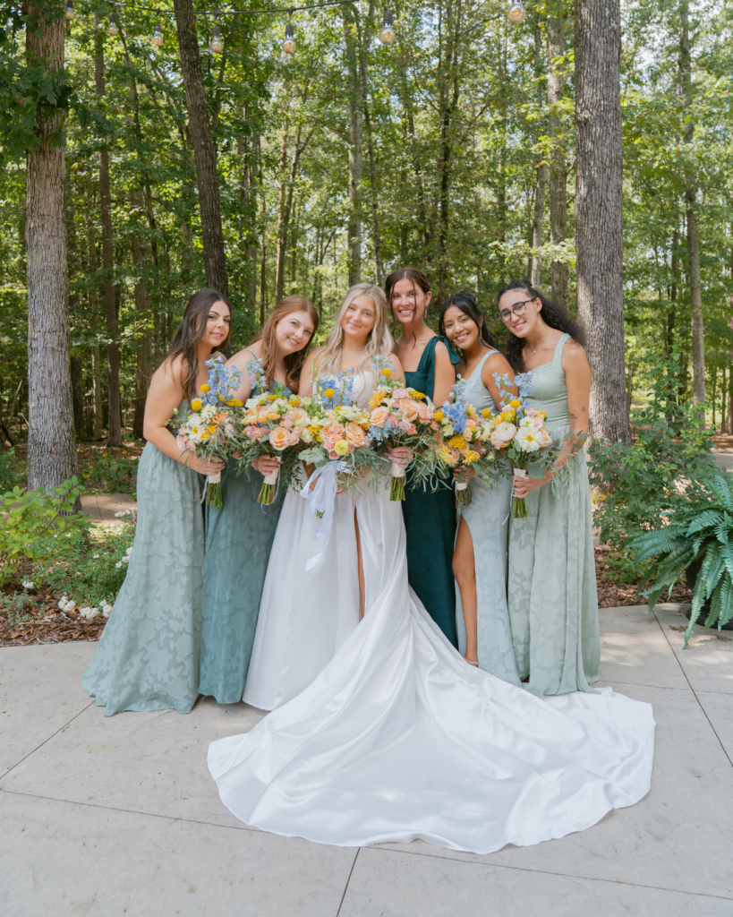 Abby poses with five bridesmaids wearing sage and deep green dresses and carrying colorful bouquets at Cedar Hills in Salisbury, NC.