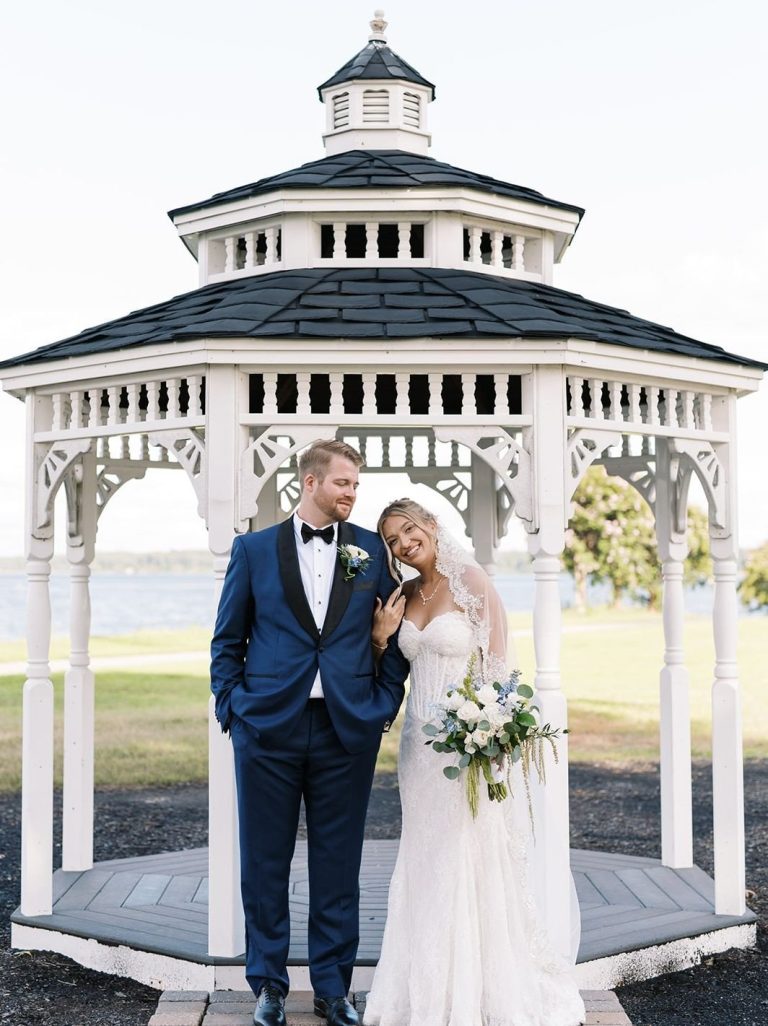 bride in a white gown and a groom in a navy blue tuxedo standing inside a white, open-sided gazebo by the waterfront.