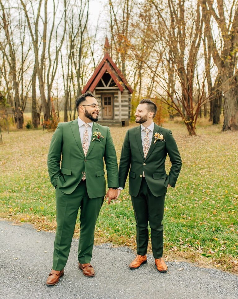 Grooms wearing green suits hold hands and smile at each other while standing in front of a small chapel surrounded by fall foliage