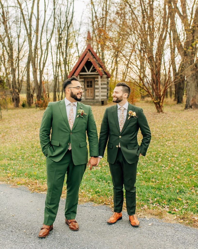 Grooms wearing green suits hold hands and smile at each other while standing in front of a small chapel surrounded by fall foliage