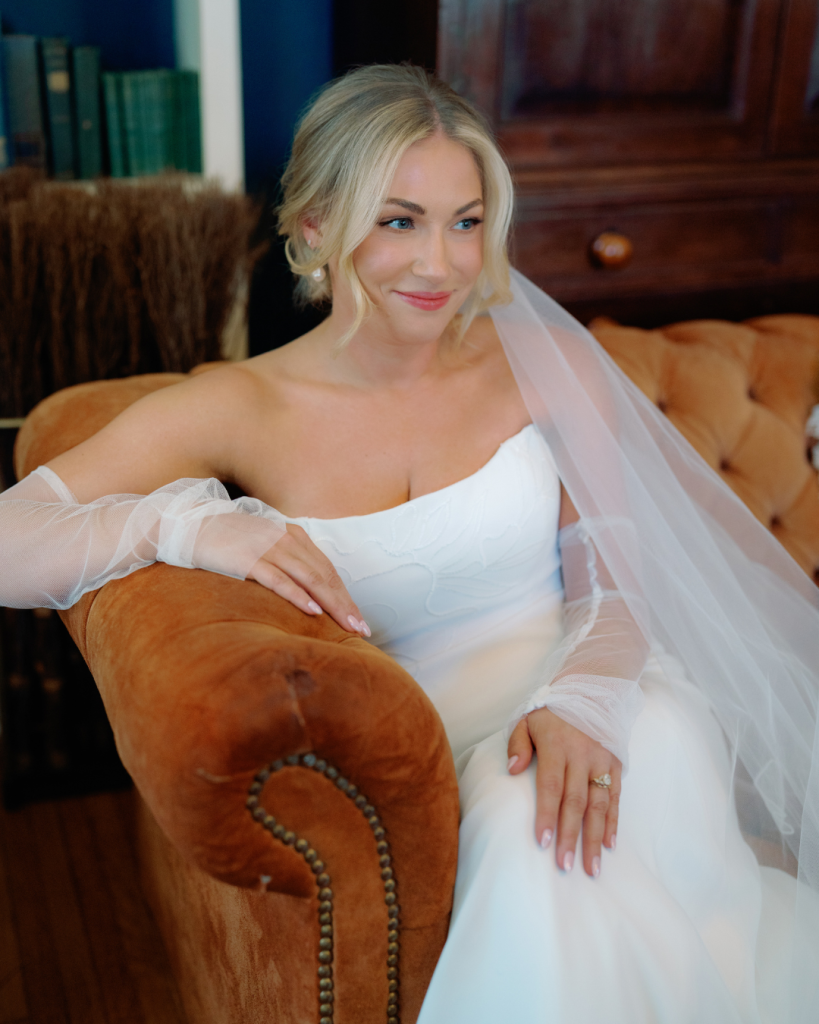 Charlotte bride Elizabeth wears a modern wedding gown with sheer sleeves and an embroidered veil while seated in a rust velvet chair.