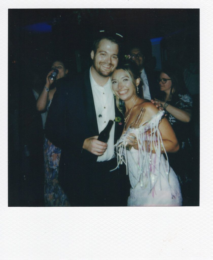 Polaroid-style photo of the bride and groom smiling together at their wedding reception, with the groom holding a beverage bottle and the bride wearing a fringed dress accessory.