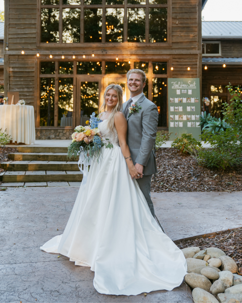 Abby and Connor pose outside the rustic Cedar Hills reception building beneath warm string lights, holding hands and smiling at the camera.