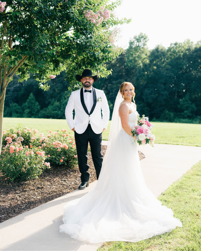 Bride and groom pose outdoors at Fields at Skycrest, with the groom wearing a white tuxedo and cowboy hat and the bride holding a pink floral bouquet.