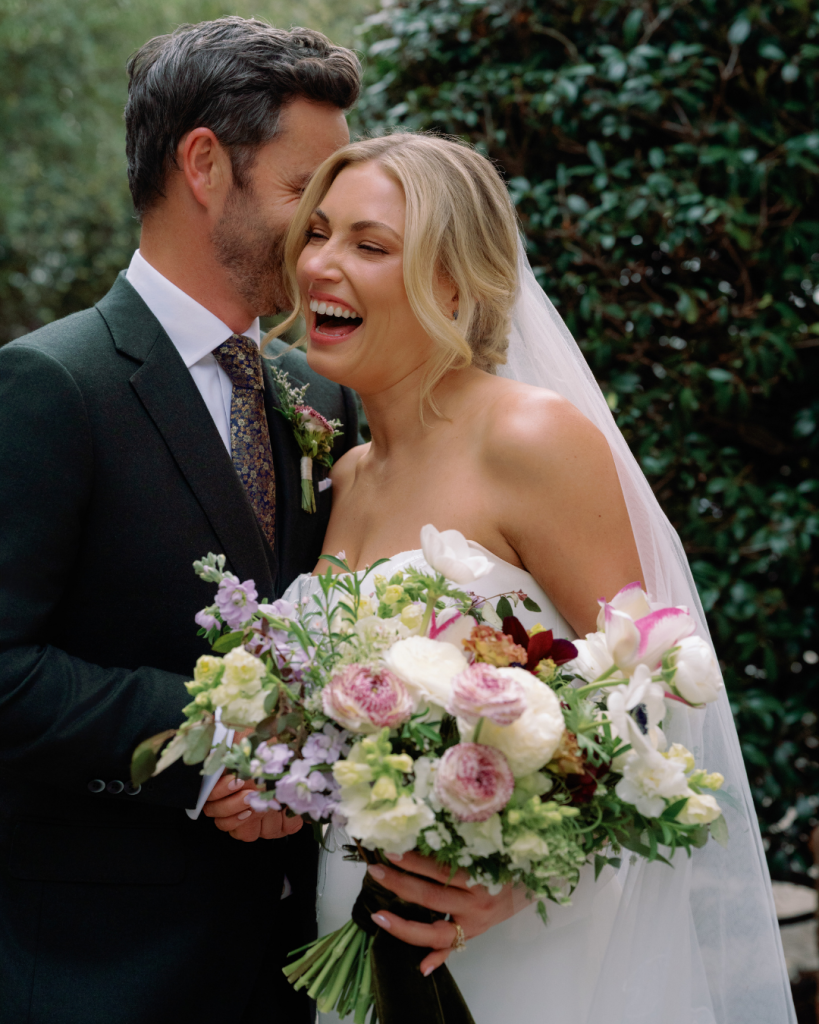 Elizabeth laughs as Tom leans close, surrounded by lush greenery and a colorful garden style bridal bouquet at The Morehead Inn.