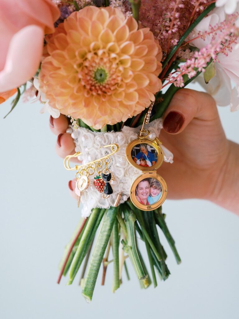 Close-up of a bride holding a bridal bouquet of large peach and light pink dahlias, with small framed family photo charms attached to the wrapped stems.