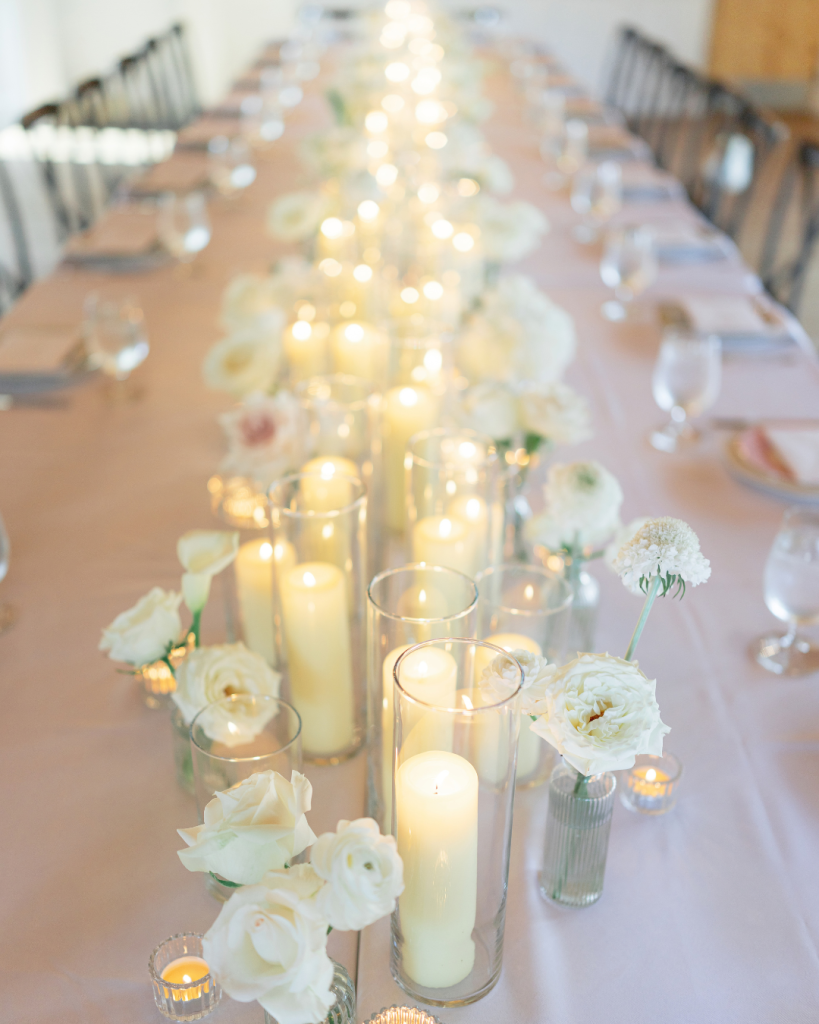 Long reception table styled with white flowers, glass vessels and glowing pillar candles for Tate and Alex’s wedding.