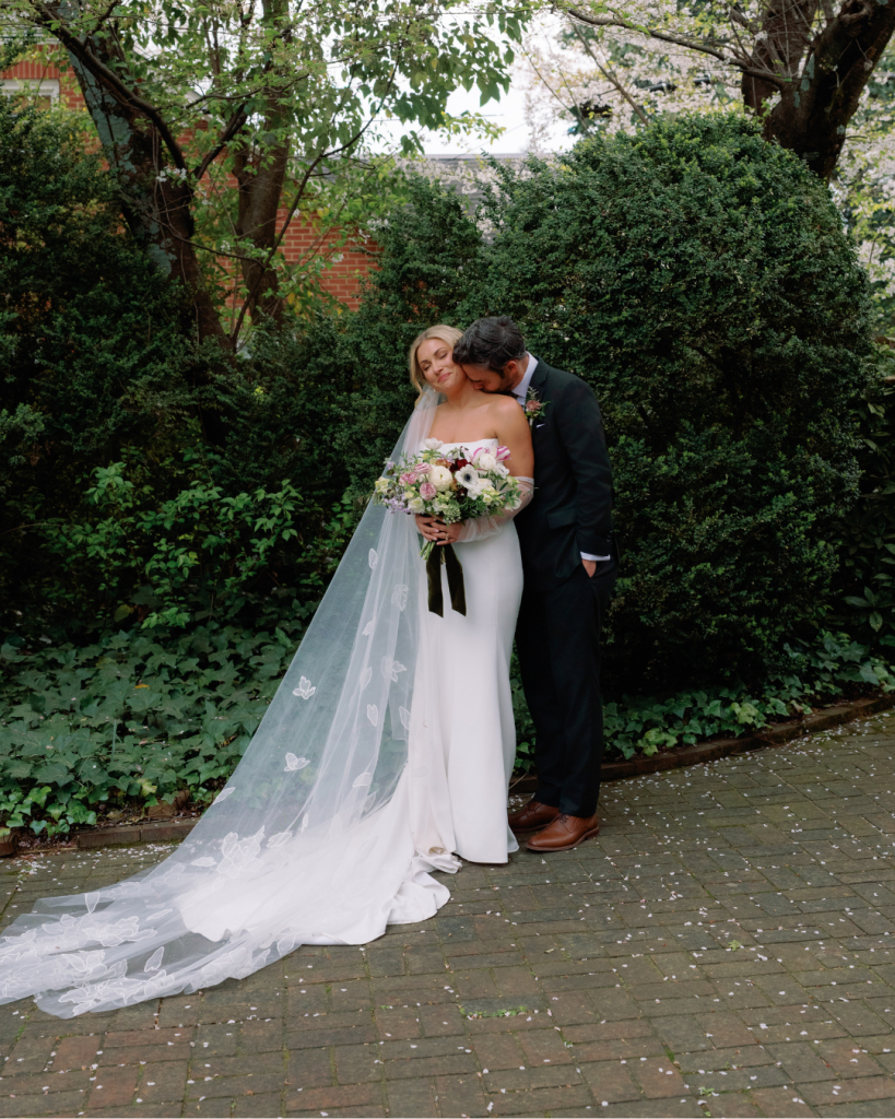 Elizabeth and Tom embrace in a garden portrait, showcasing the bride's long embroidered veil and romantic bouquet.