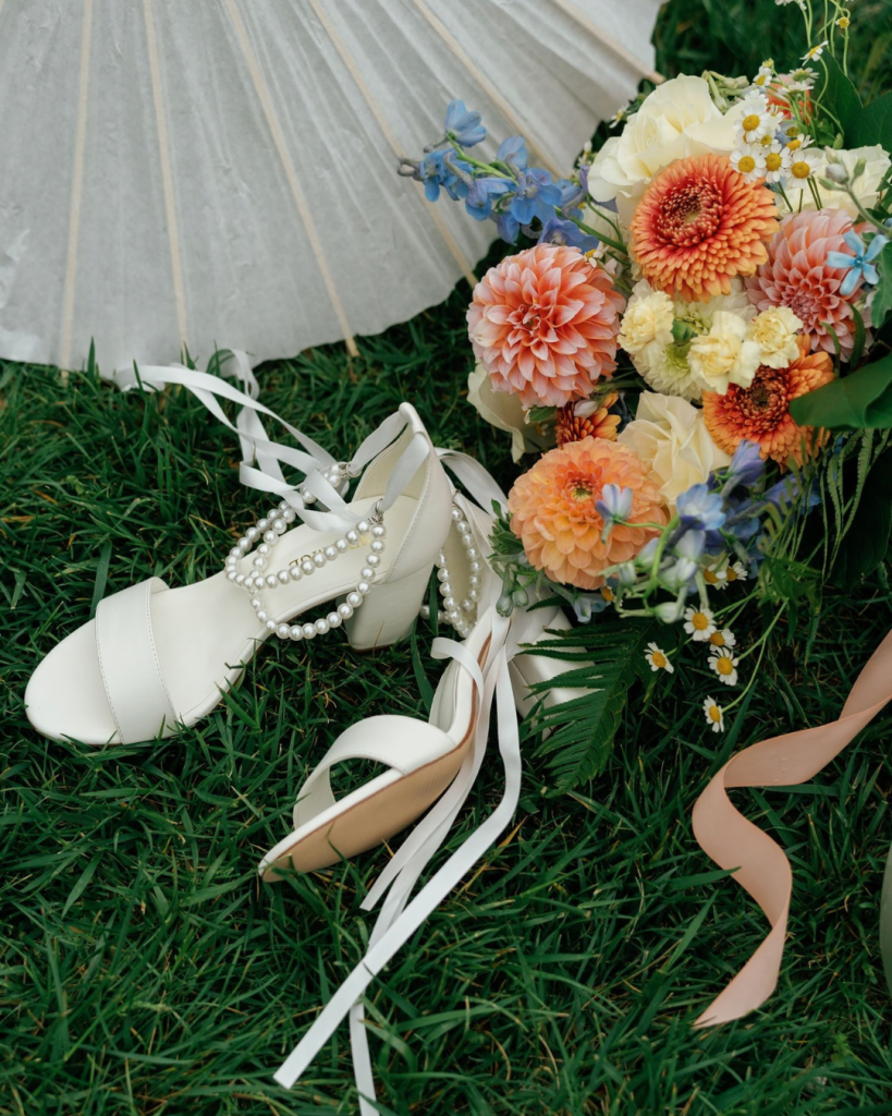 White bridal heels with pearl straps and ribbon ties rest beside a colorful spring wedding bouquet featuring peach dahlias, yellow roses, blue flowers, and delicate daisies.