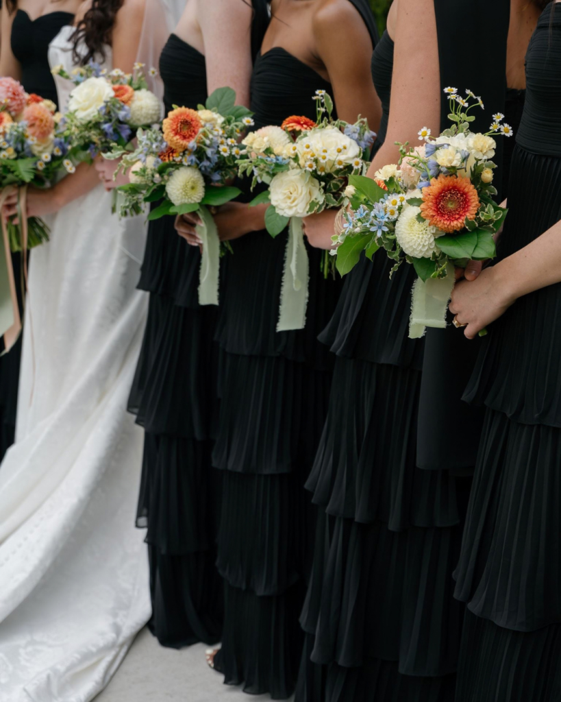 Bridesmaids in black tiered dresses hold colorful bouquets of peach, coral, yellow, white, and blue spring flowers at White Willow Meadows.