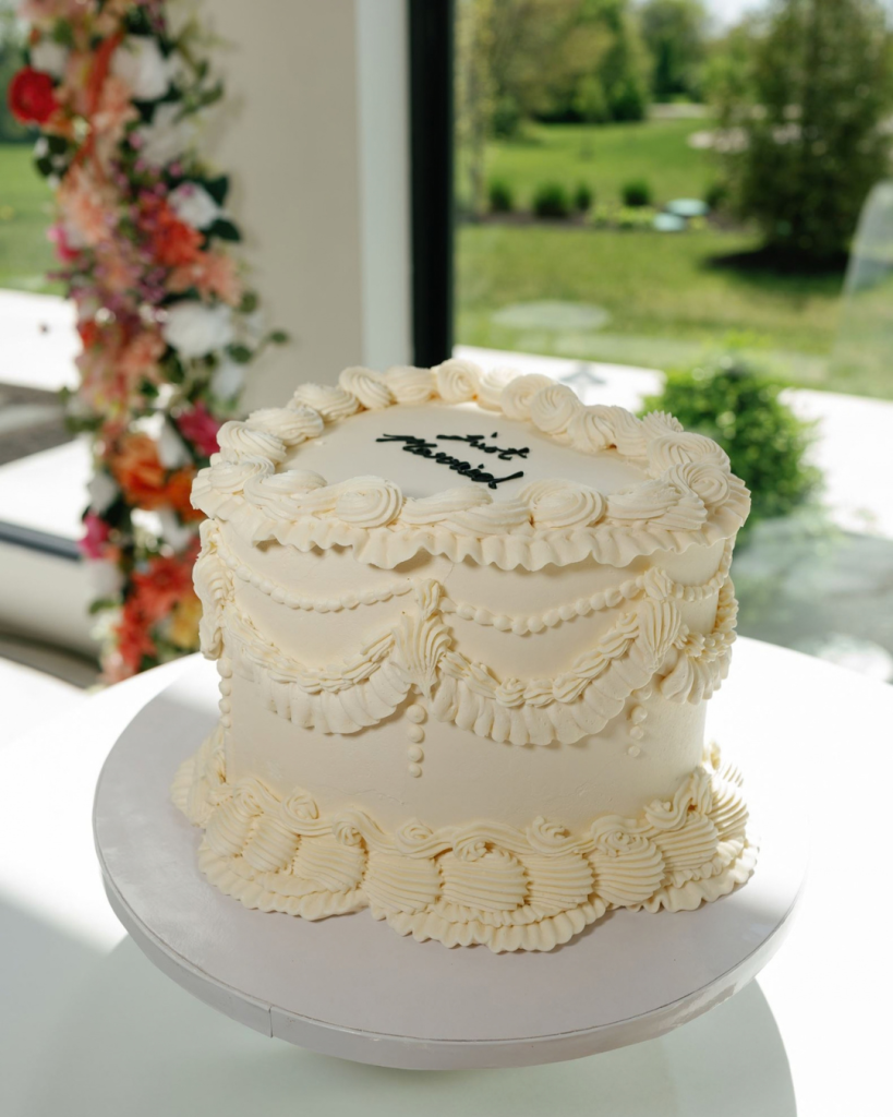 Vintage-inspired white wedding cake featuring intricate piped frosting, decorative swags, and a scalloped border at Charis and Jotham's spring wedding.