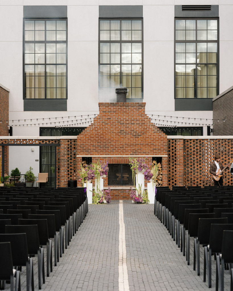 Outdoor wedding ceremony courtyard at The Junto in Columbus with black chairs, brick fireplace, and purple floral installations.