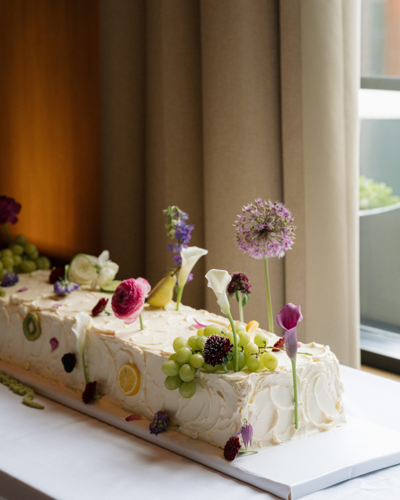 Long white wedding cake decorated with fresh flowers, grapes, and citrus slices at a spring wedding at The Junto in Columbus