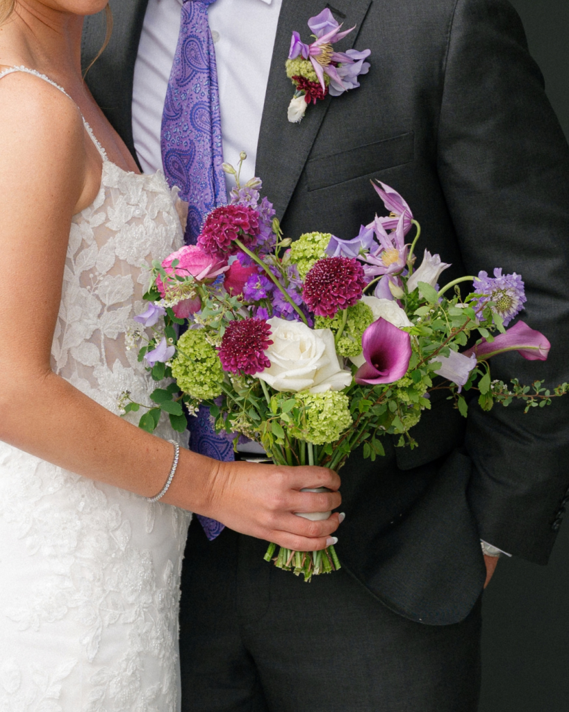 Close-up of bride's colorful purple, pink, and green wedding bouquet beside groom's charcoal suit and purple patterned tie